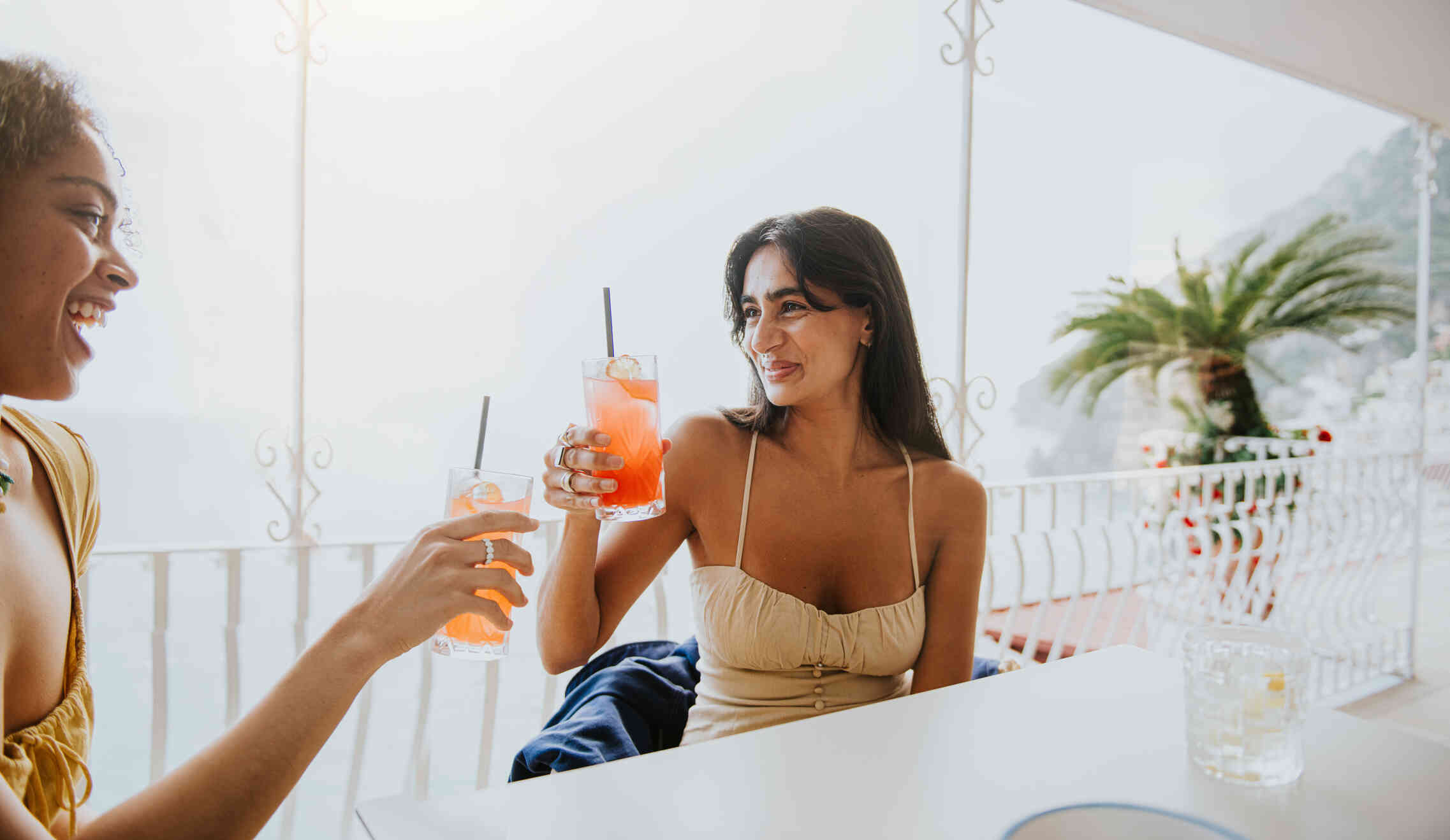 Two woman sit next to each other at a table and lift their cups filled with a colorful drink while smiling.